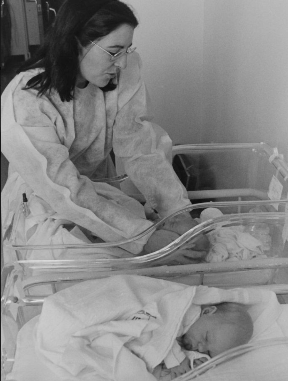 A woman in hospital garb tends to two babies in clear, hospital cribs.