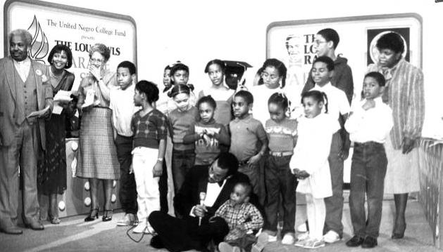 A group of young children stand together. An adult sits on the floor with one child and holds a microphone. 