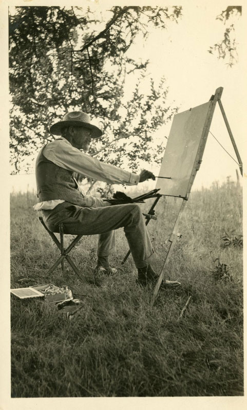 A man is sitting in a chair in the middle of an open field. He is painting on a canvas set up on an easel.