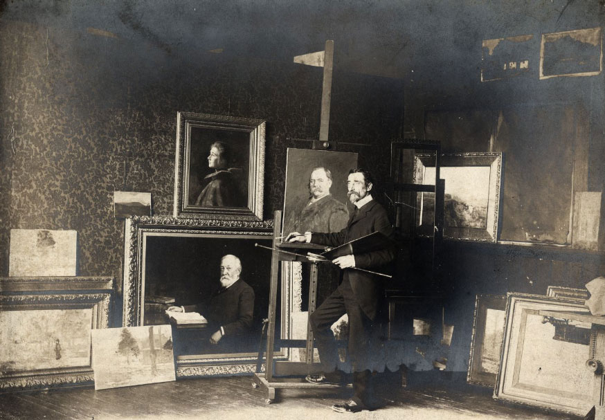 Steele stands holding painting tools. The room is filled with his completed paintings. 