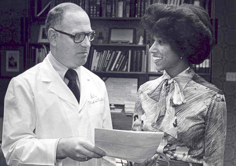 A man in lab coat holds a document as if showing it to the woman beside him.