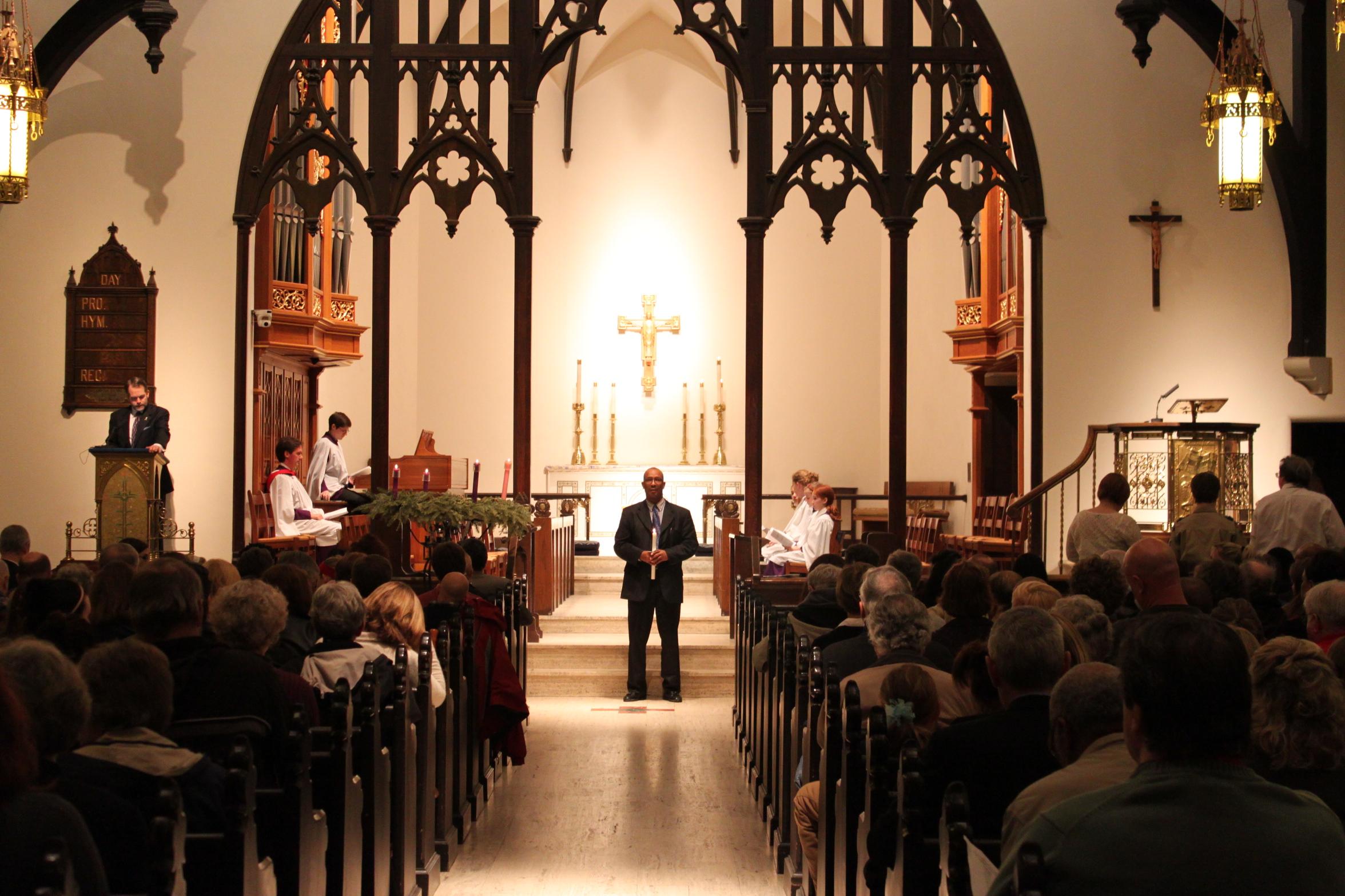 People fill the pews of a church. A man stands at the front holding a candle.