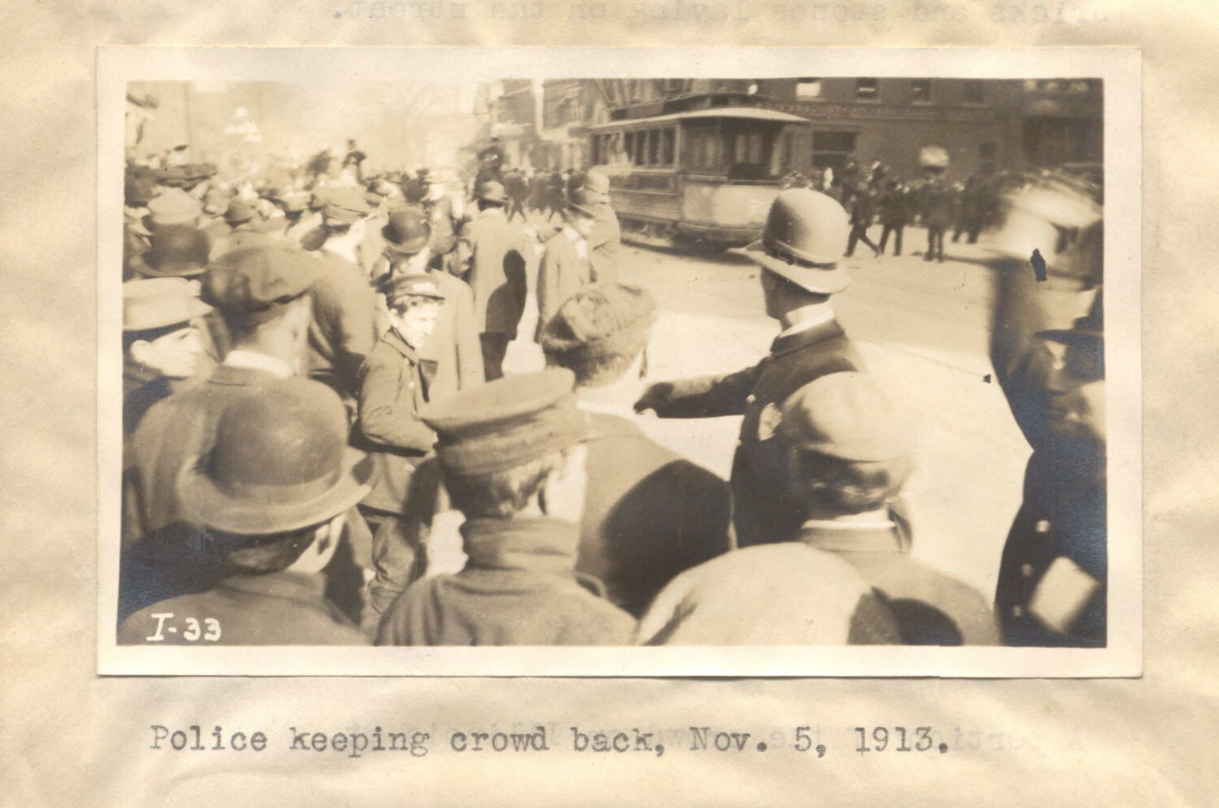 A police officer holds back a crowd. A streetcar runs down the road in the background.
