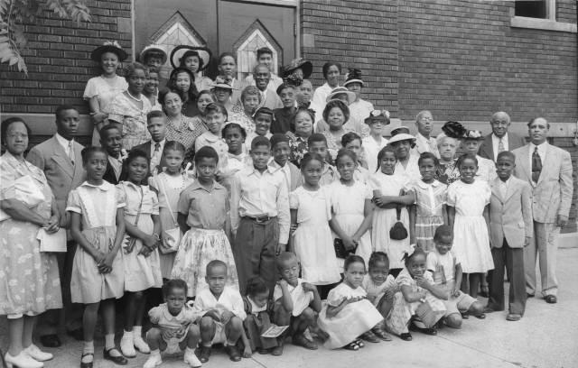 A group of children and several adults are gathered outside of a building for a group photograph.