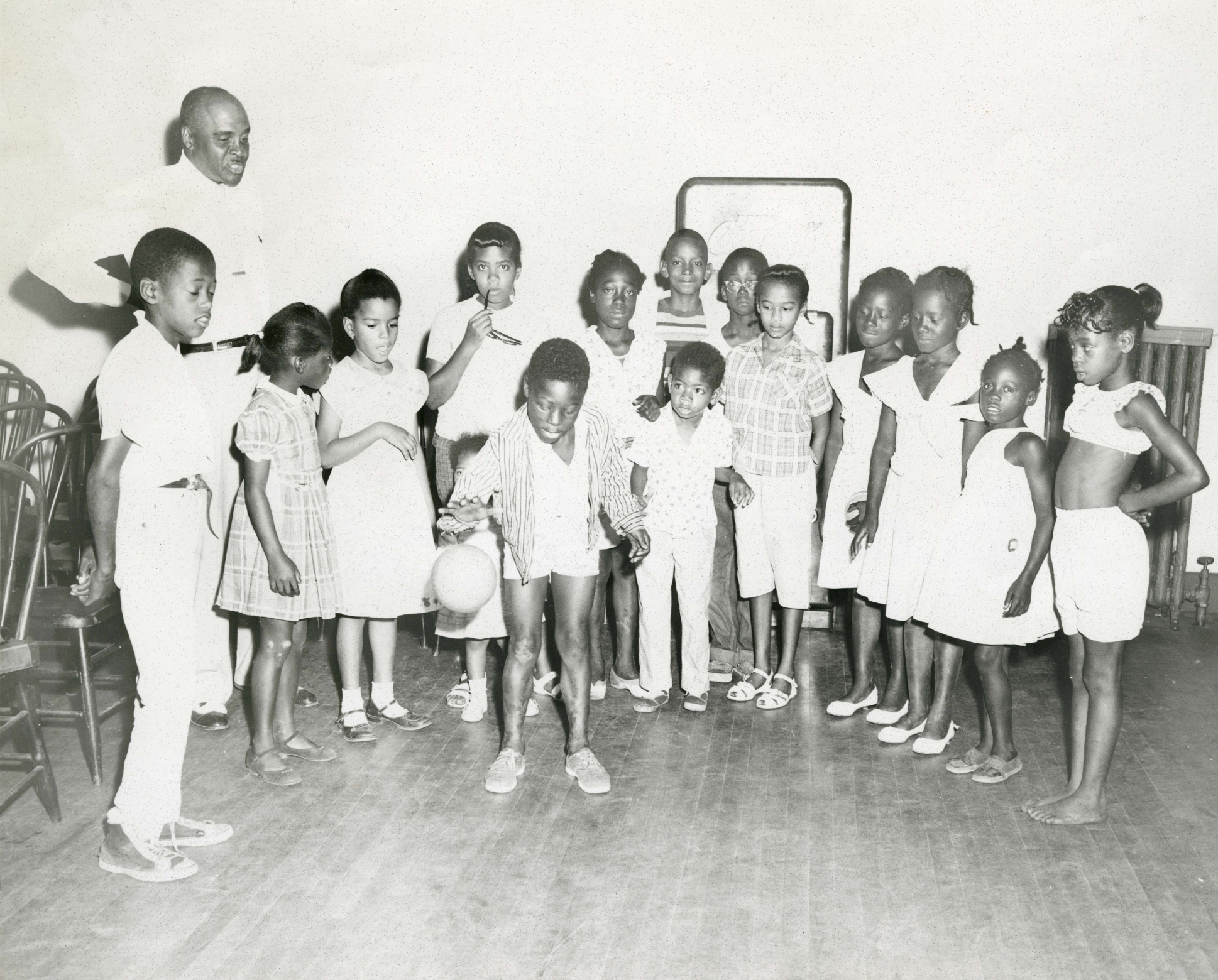 A group of children and one adult are gathered around a young boy and watching him bounce a ball.