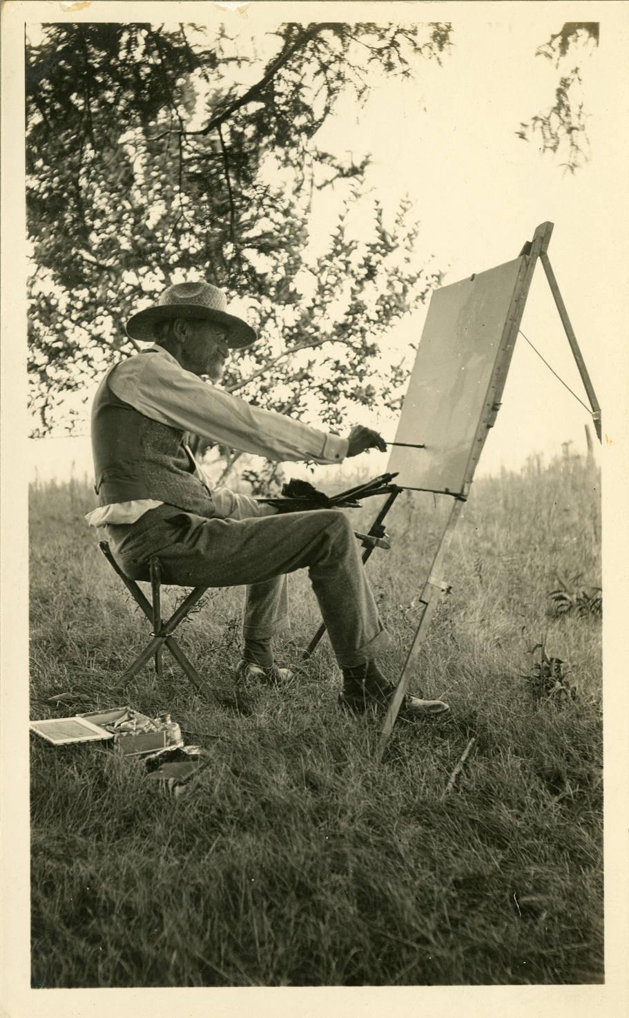 A man is sitting in a chair in the middle of an open field. He is painting on a canvas set up on an easel. 