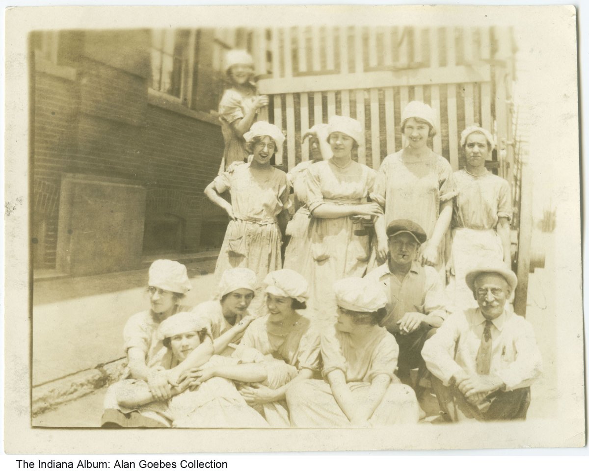 A group of young women in smocks and bonnets and two men pose together for a group photo outside of a factory building.  