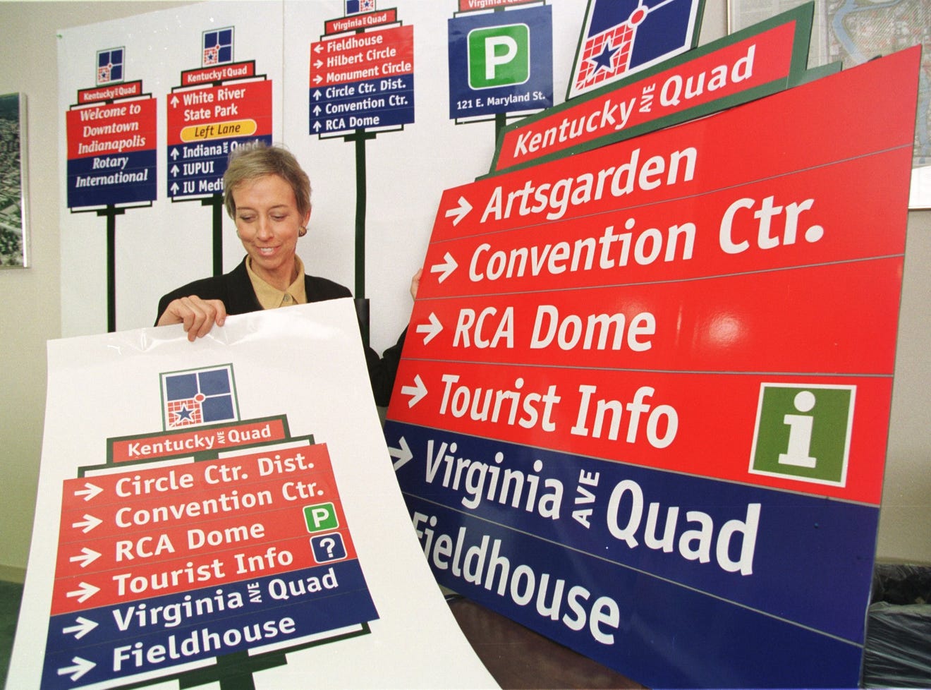 A woman holds up a poster showing a design of a sign with arrows next to location names. More designs are in the background. 