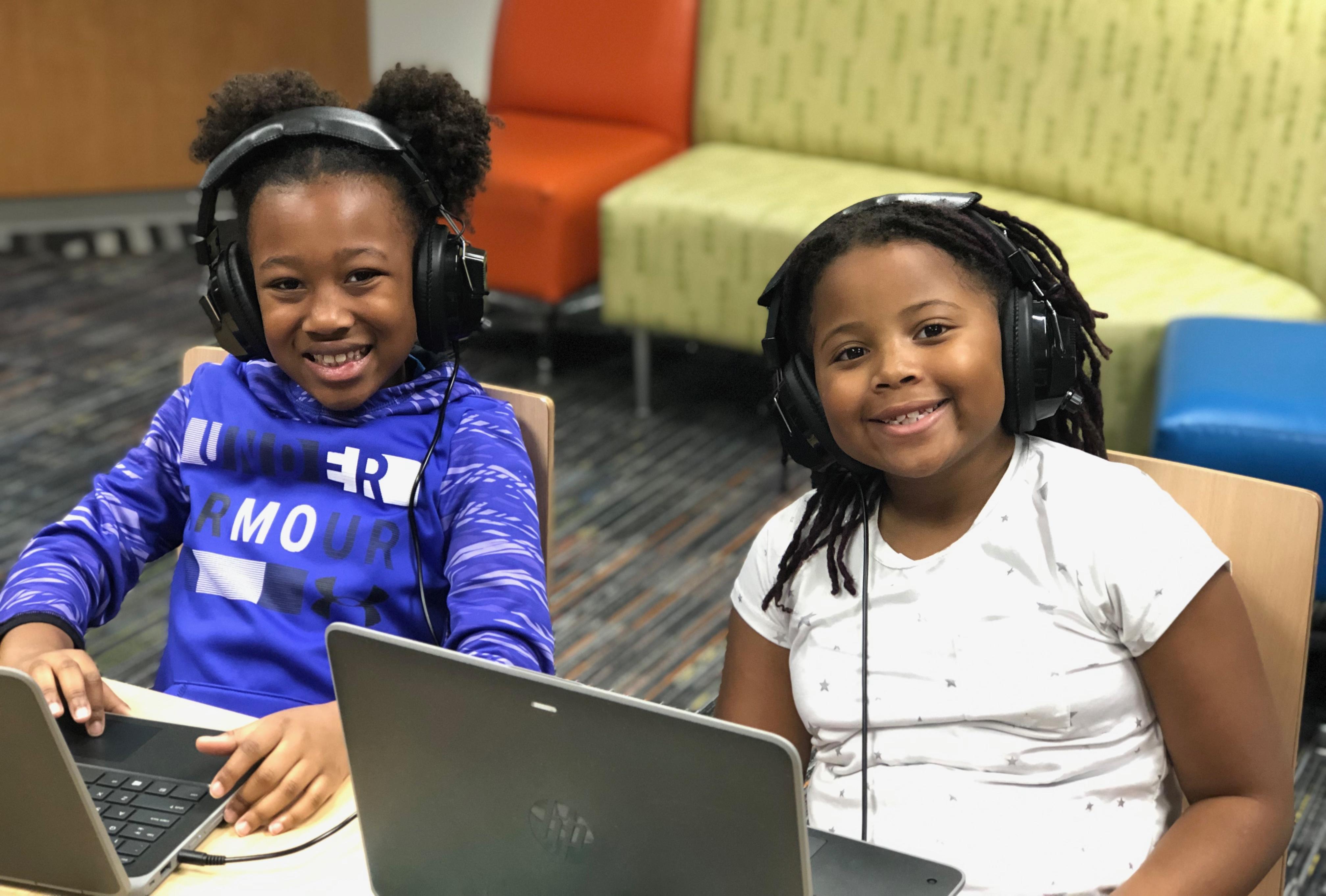 Two young children sit in front of laptops and are wearing headphones. 