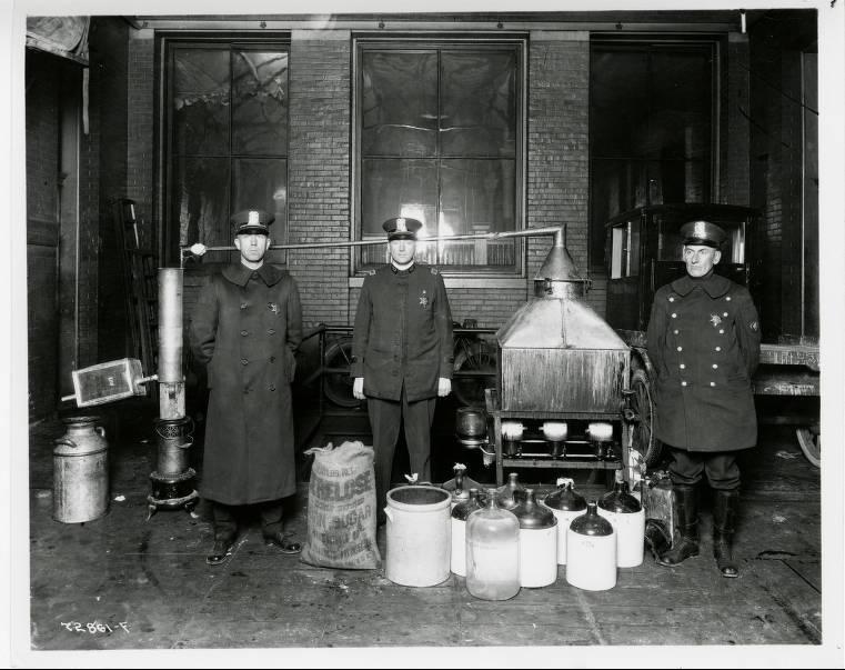 Three police officers stand with jugs of alcohol.
