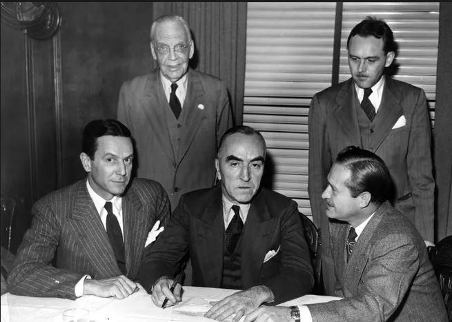 Five men sit and stand around a desk.