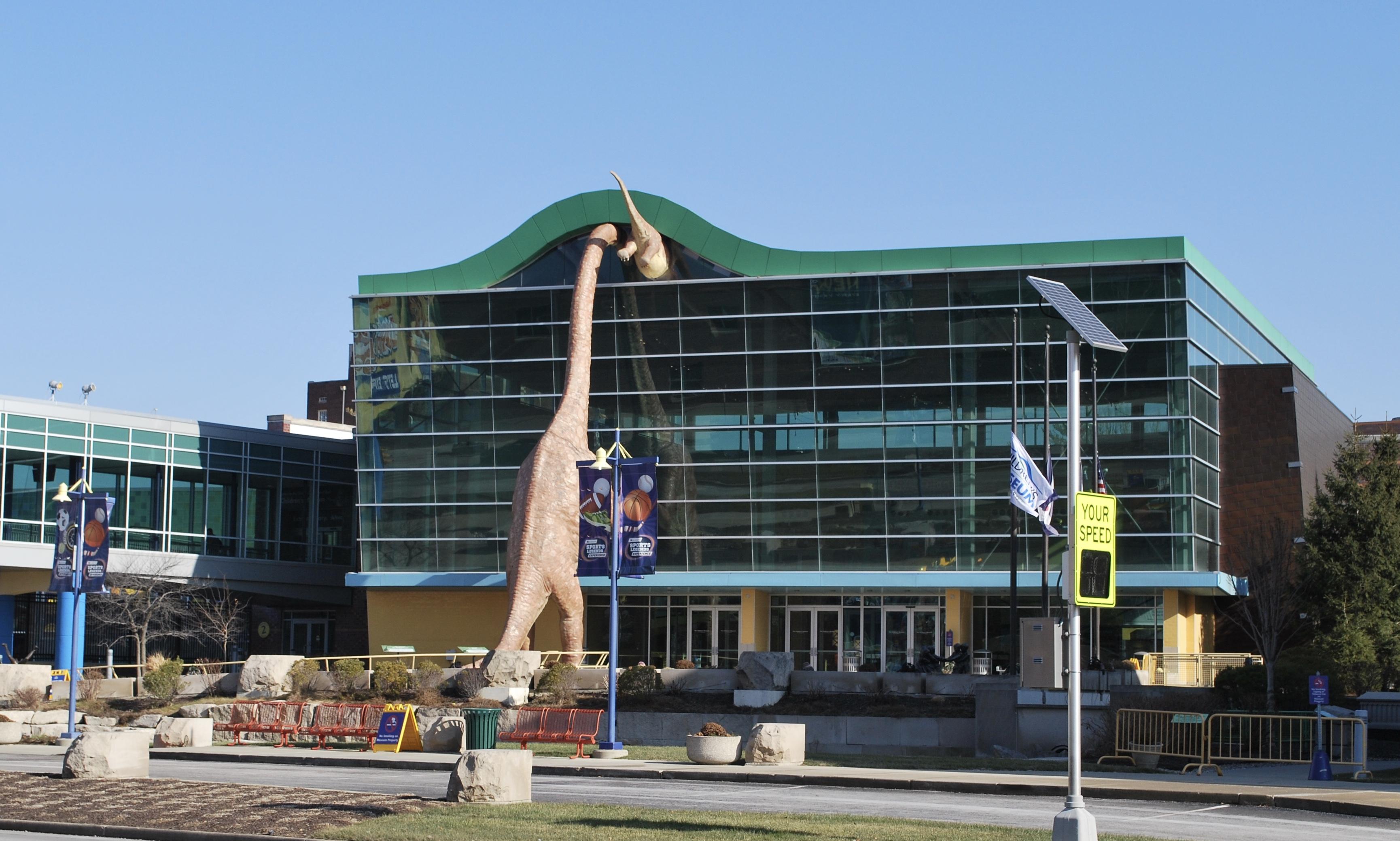 A dinosaur statue that is as tall as the building is situated so that it appears to be looking into the glass front. 