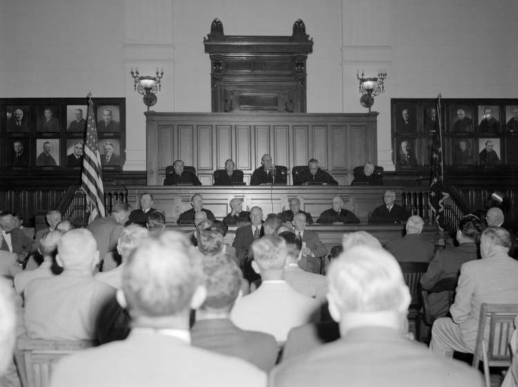 Two rows of judges sit at the front of a room. The audience can be seen in the foreground.