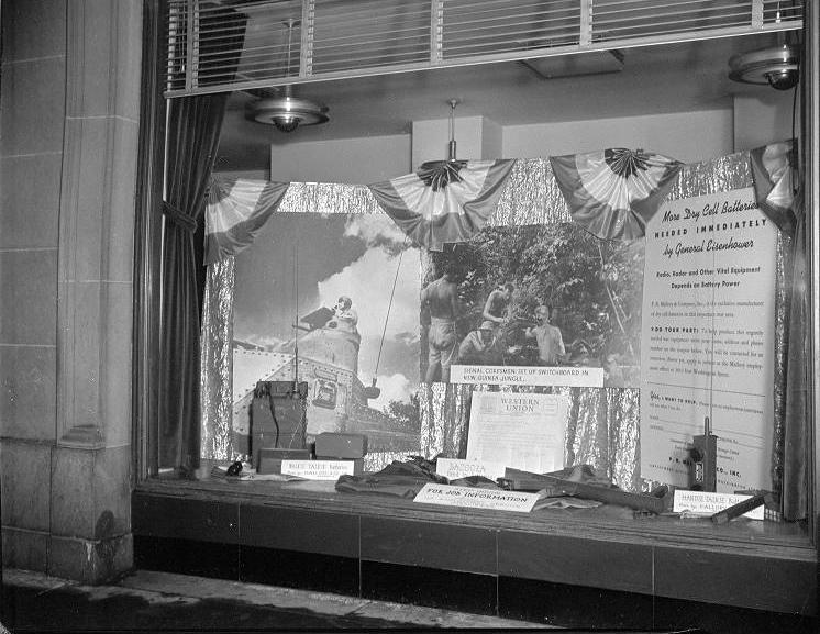 A window display featuring images of men using equipment powered by P R Mallory's batteries as well as physical examples of the equipment. 