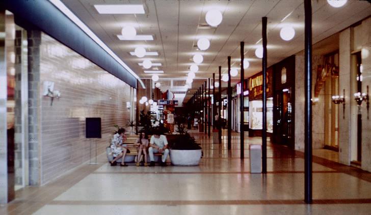 Three people sit on a bench in the middle of a mall corridor. 