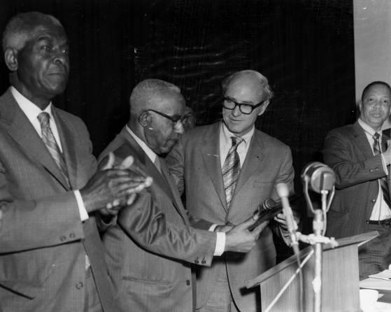 Four men are standing in a row behind a podium and microphone.