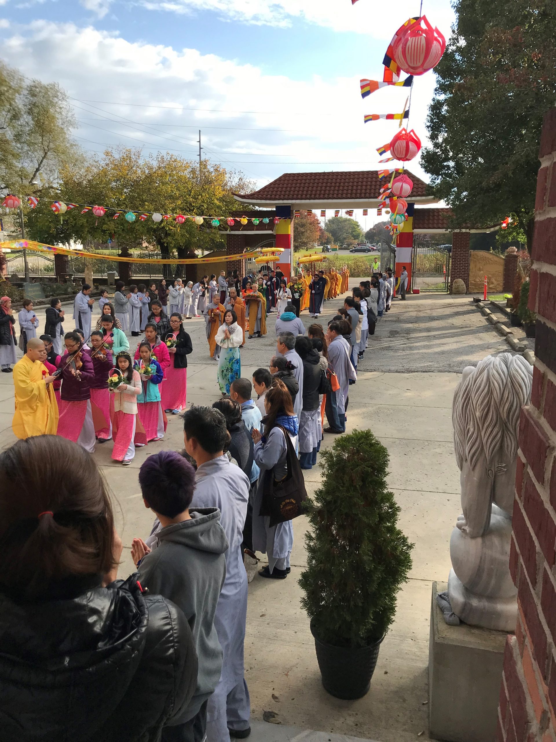 Two rows of people, most in gray robes, line the procession on a long, concrete courtyard. The yellow-robed monks, proceeded by colorfully-dressed women holding flowers, process from a pagoda archway at the far end.