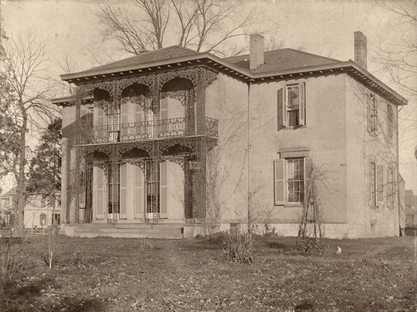 Exterior view of a two story home with ornate iron work around the lower and upper parts of the front porch. 