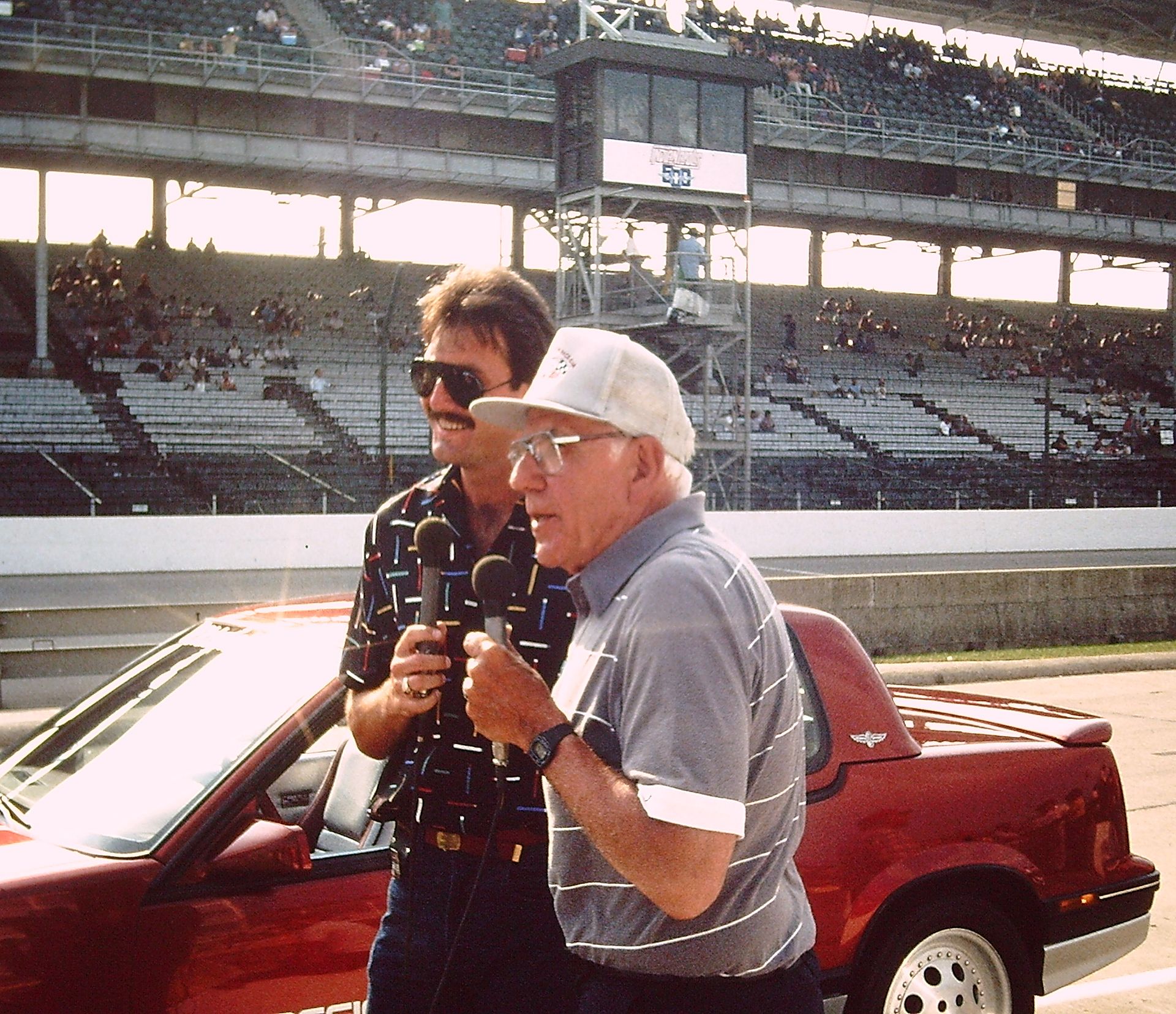 Two men holding microphones stand next to a pace car in Pit Row at the Indianapolis 500 Racetrack.