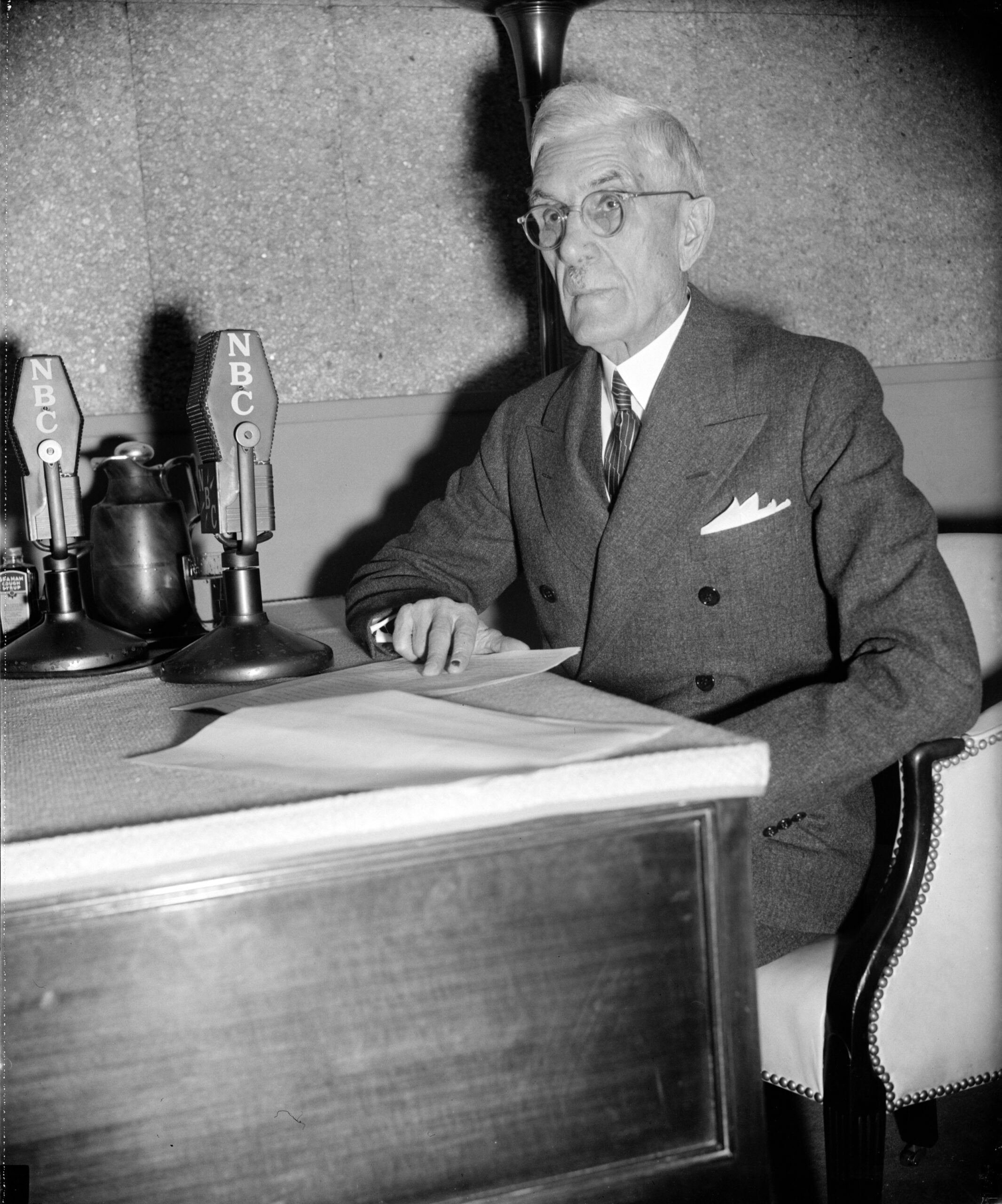 Dr. Francis Townsend sits at a desk reading off papers into microphones. 