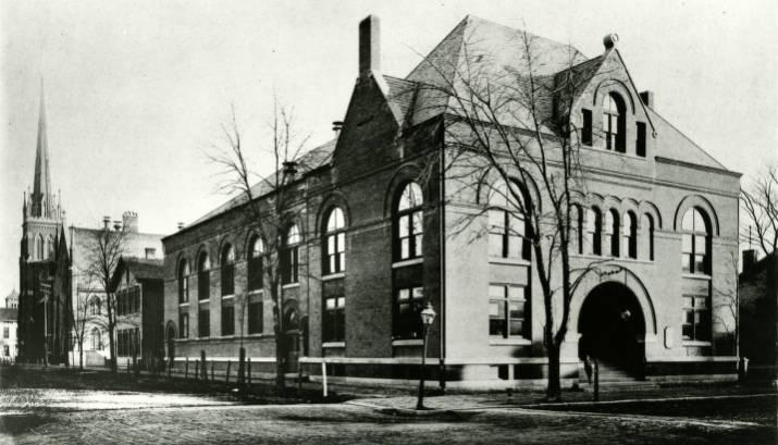 The long, two-story brick building has a hipped roof with a front and side gable, large arch windows on the second floor and an arched entrance.
