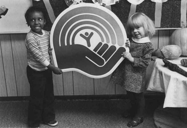Two children hold a sign with the United Way logo on it. The logo is a hand with a human-like figure and a rainbow at the top. 