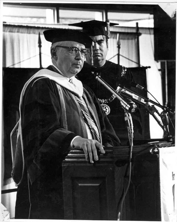 Two men stand speaking at a podium. They are both wearing academic regalia.
