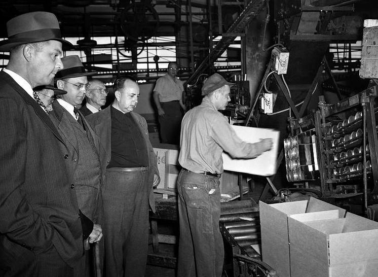 A worker is lifting a box off of a factory line while a group of men watch.