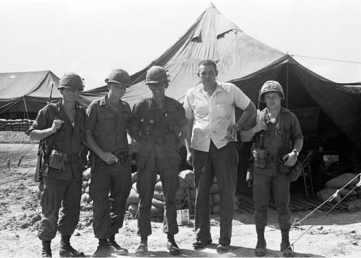 A man in civilian dress stand with four soldiers with tents in the background.