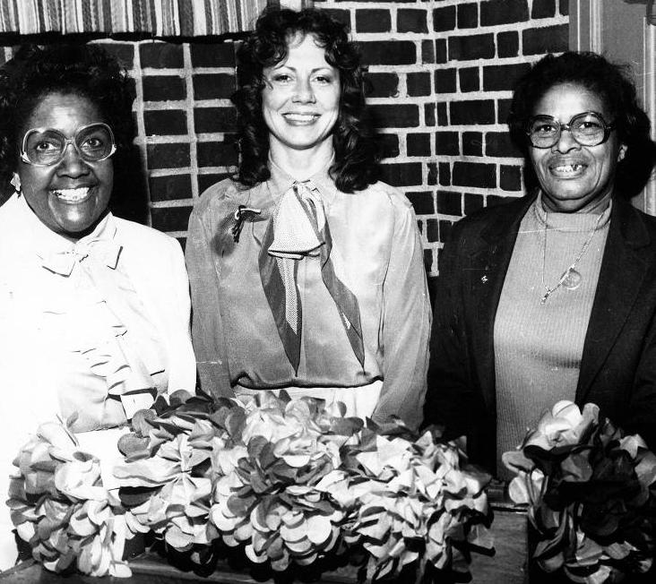 Three woman stand together with flowers sitting on the table in front of them.