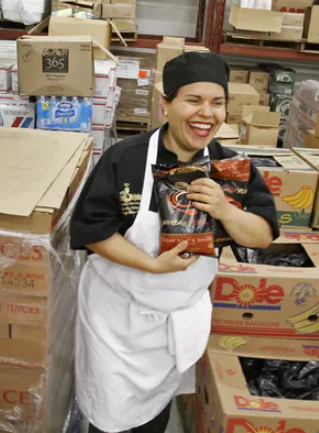 A smiling woman holds bags of food. Boxes of groceries fill the background.