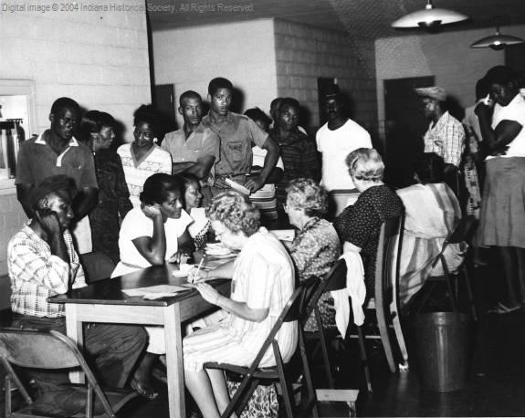 A row of people sit at a table. People sit opposite them while others stand in a line.
