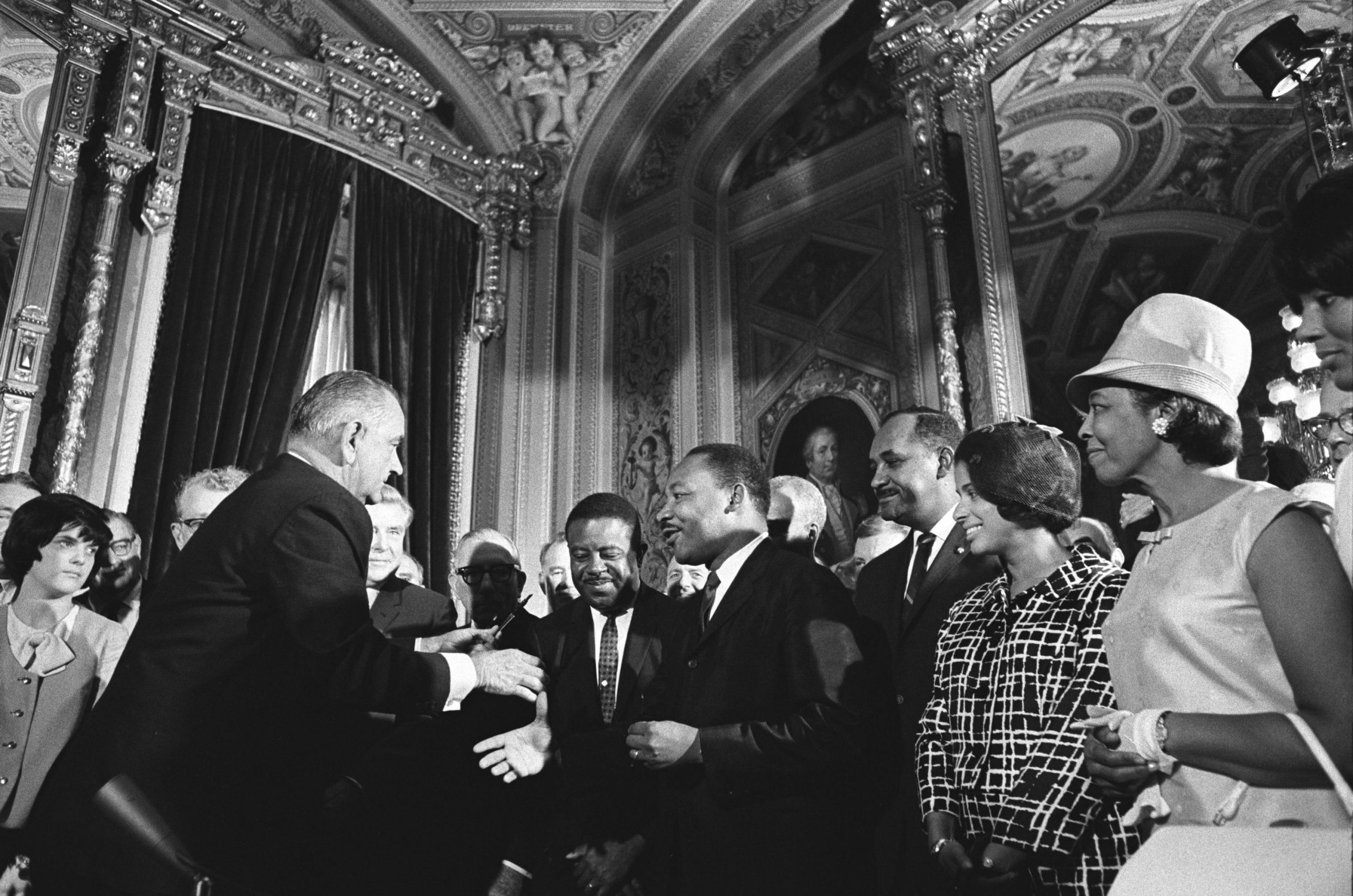 President Johnson and Martin Luther King Jr. shake hands. Other people stand around them watching.