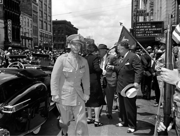 A man in a military uniform stands in the middle of a crowded city street. 