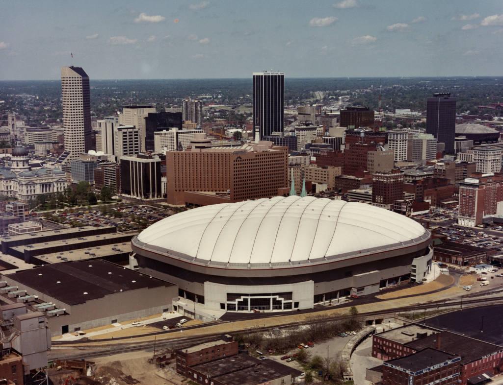 A large, domed arena sits front and center in an aerial view of downtown Indianapolis. Beyond it are the state capitol building and the many other office buildings.