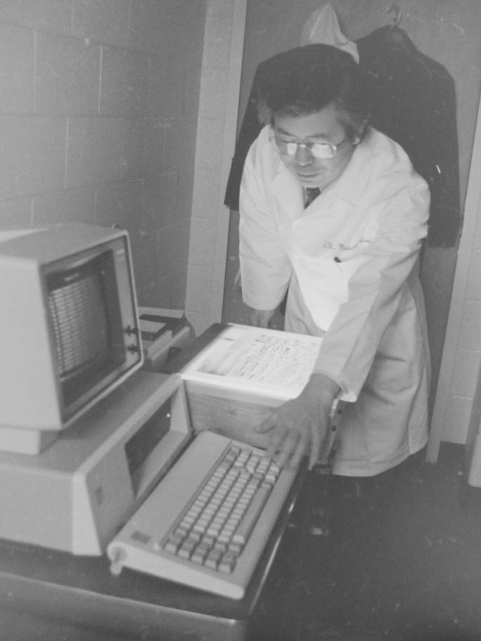 A man in a lab coat gestures towards a computer. 
