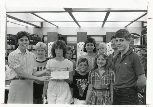 An adult presents several children with a certificate. Library shelves full of books are in the background. 
