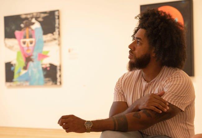 A man sits in the foreground. Two pieces of art are hanging on the wall in the background.