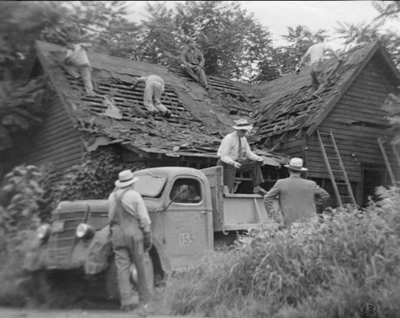 A truck is parked outside of a dilapidated house. Several men are working on the roof.