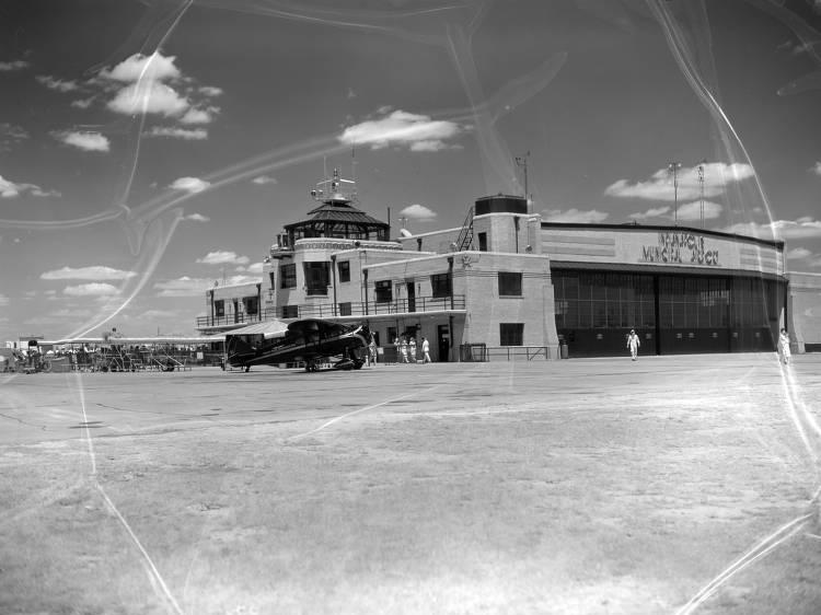 A flat-roofed, two-story building with a glass front sits on a large concrete area with a small plane out front.