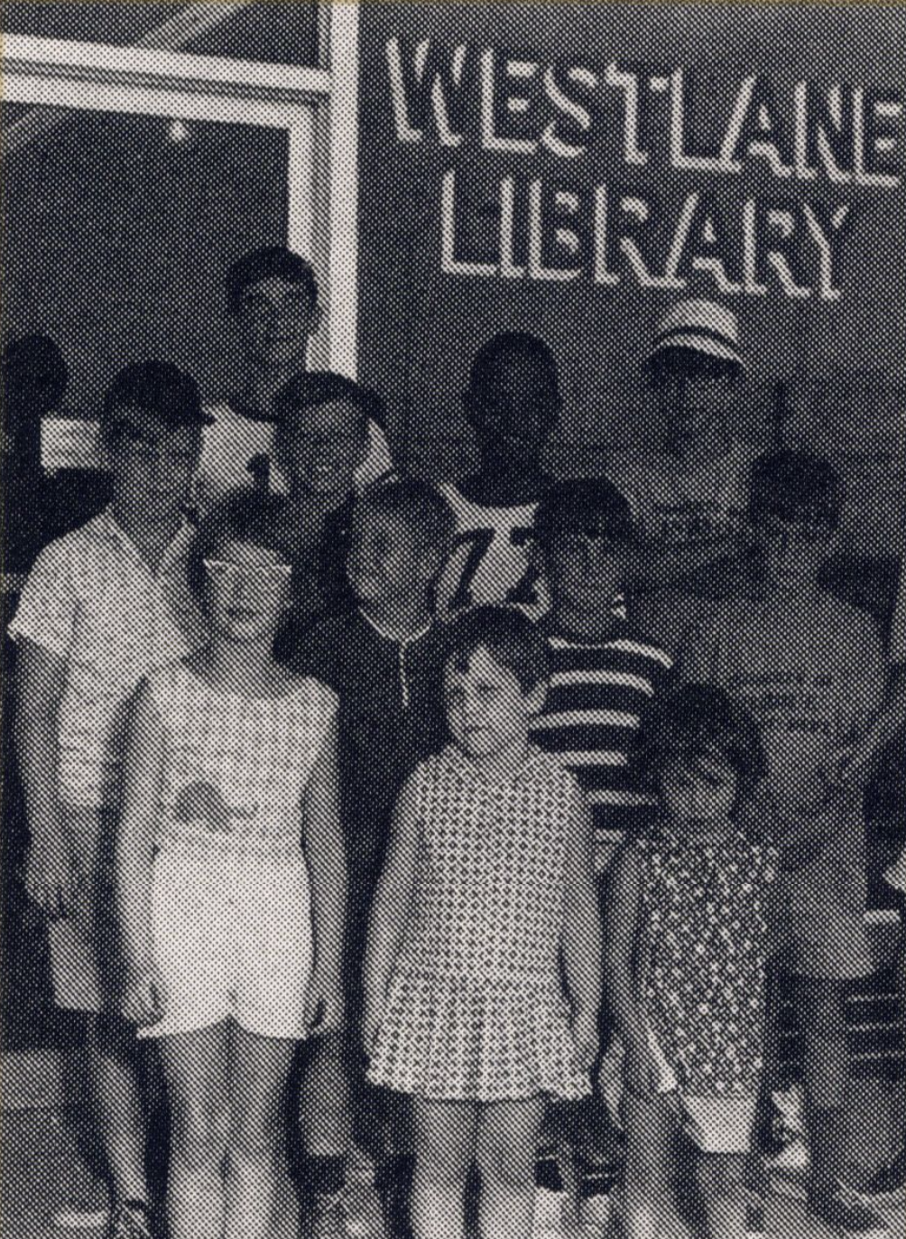 A group of children stand outside of the entrance to a building. A sign on the building reads "Westlane Library."
