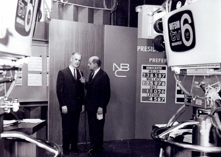 Two men talk on a television studio set. 