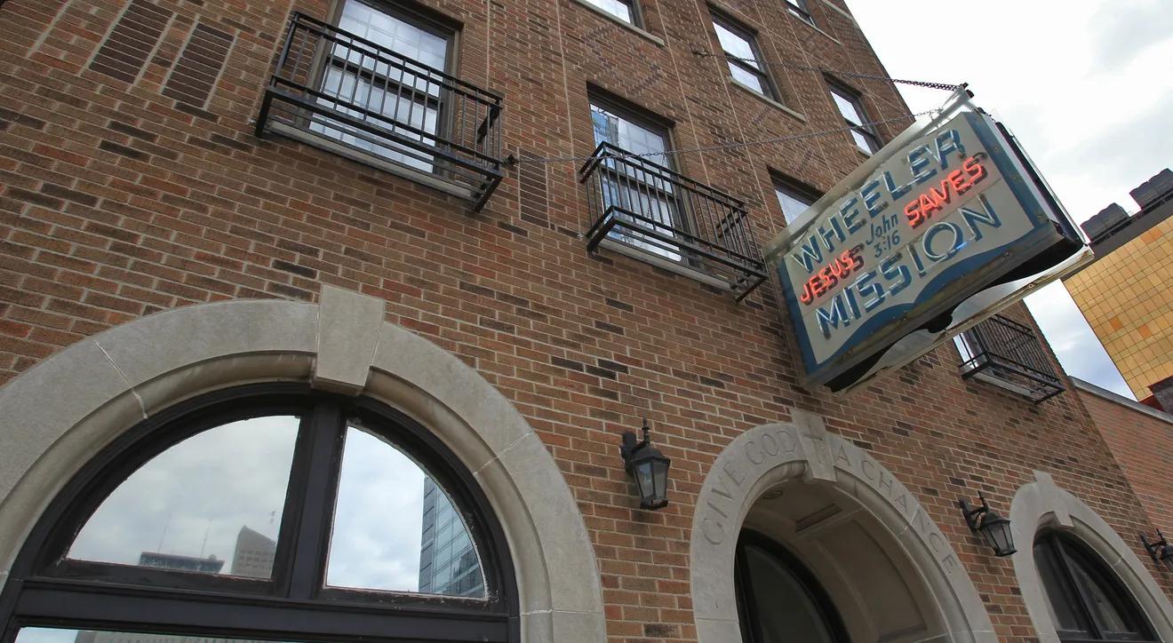 Close up of a brick building with a sign hanging off the front that reads "Wheeler Mission."