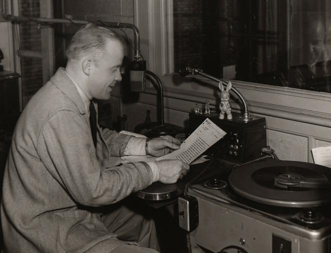 A man sits on a stool in front of microphones and turn tables. He is reading off of a piece of paper.