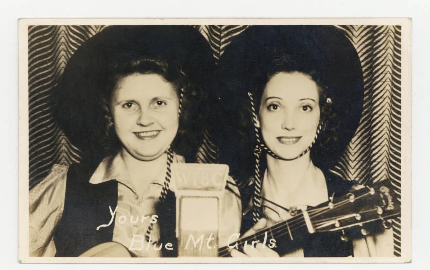 Two women stand in front of a microphone. They are wearing western-style hats and one woman is holding a guitar.