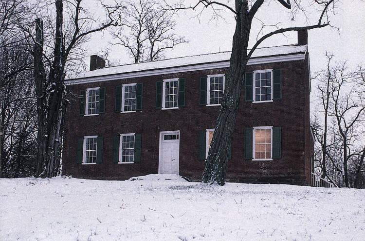 A brick two-story Federal style building has a peaked roof with a chimney at each end white-trimmed windows and a white front door.
