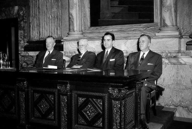 Four men sit at a desk next to each other.