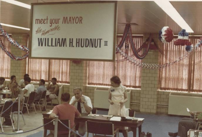 Mayor Hudnut sits at a table and chats with a man. A woman is standing next to them. A large sign hangs above them that says "Meet your Mayor."