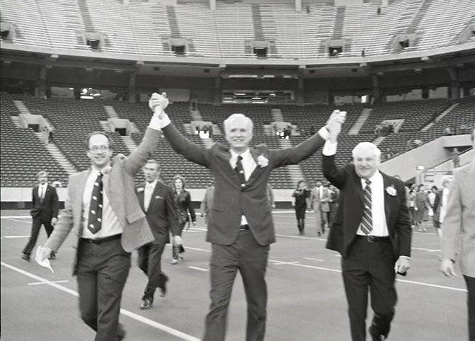 Mayor Hudnut stands between two men on a football field. He is holding up each of their hands in celebration. 