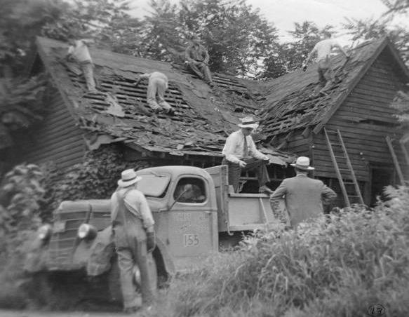 Four men are tearing the roof off of a dilapidated house. There are three men and a truck in front of the house.
